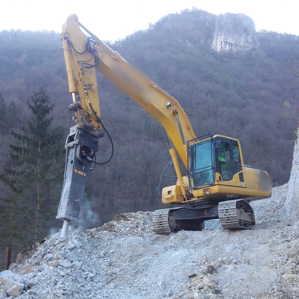 Italdem GK series hydraulic breaker mounted on a yellow excavator, breaking rock in a mountain quarry